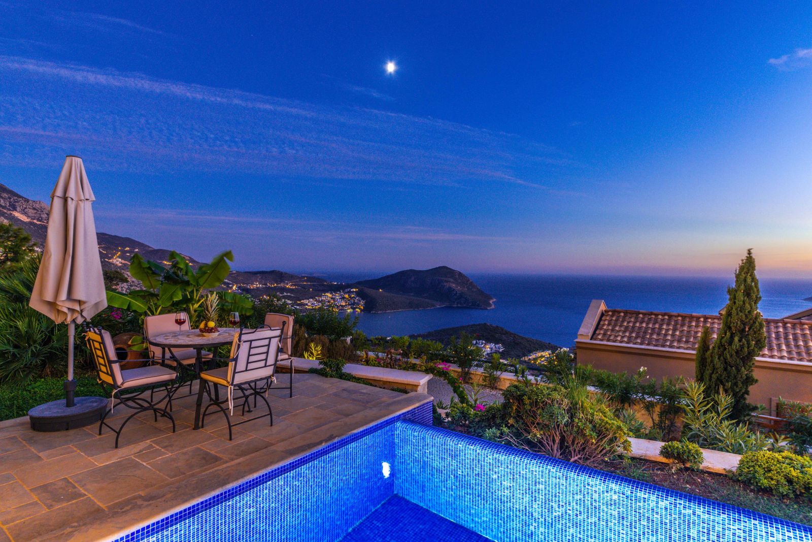 A terrace with a table and parasol above a pool at dusk, looking over the sea and the hills