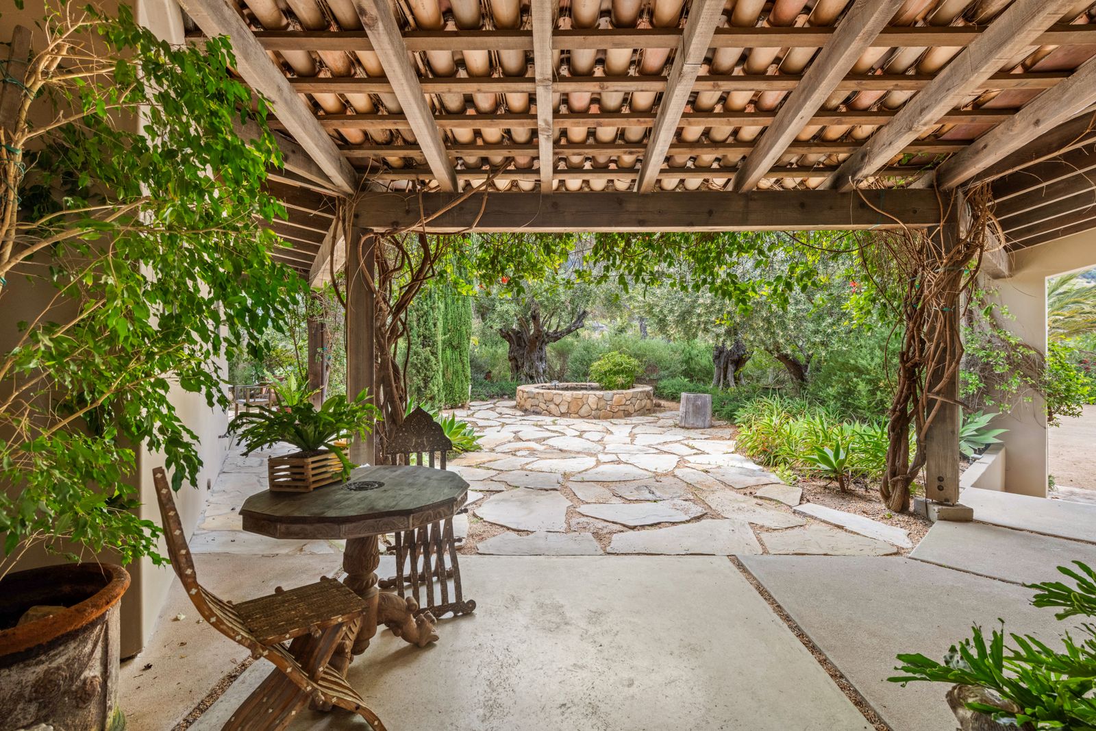 A shaded stone patio under a timber pergola, with a small table and a garden path