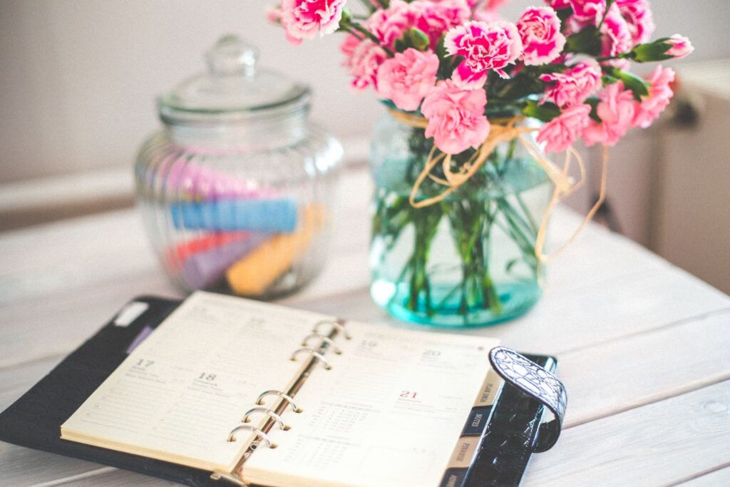 An open planner beside a vase of pink flowers on a desk