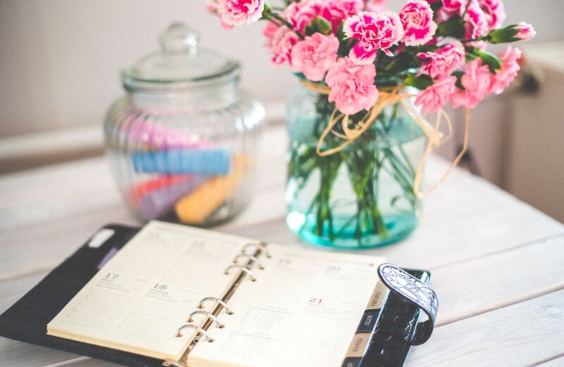 An open planner beside a vase of pink flowers on a desk