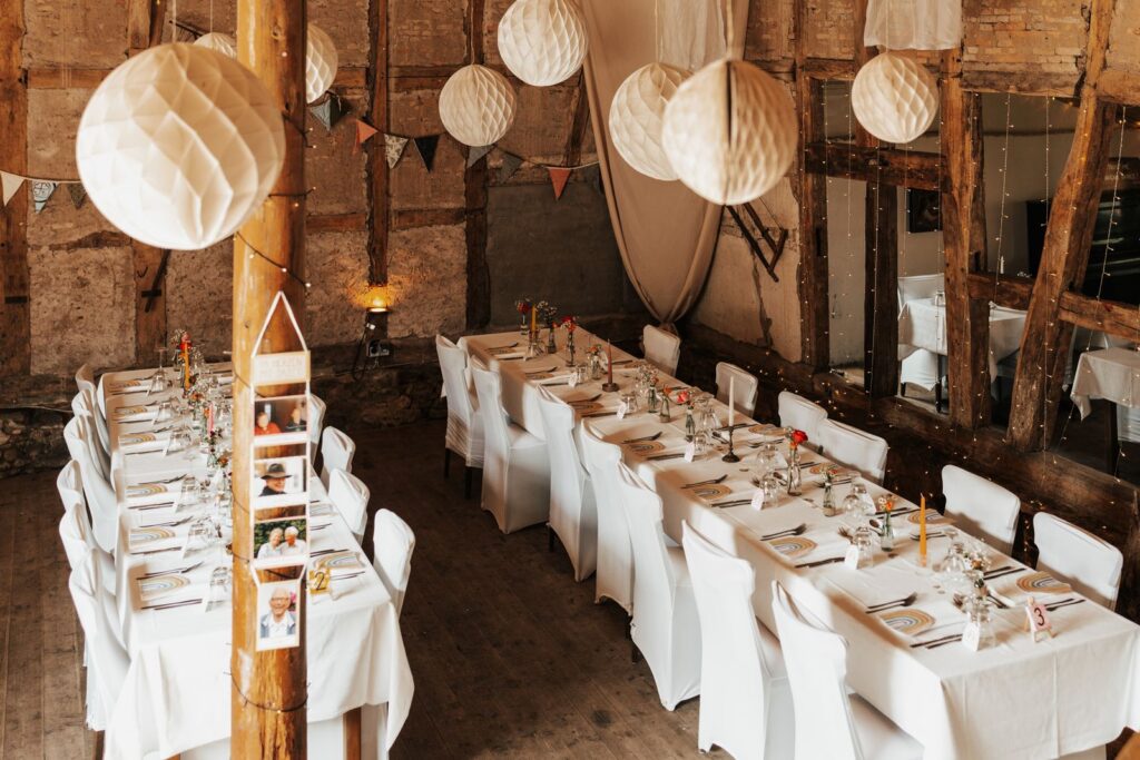 Long reception tables with white chairs under paper lanterns in a barn