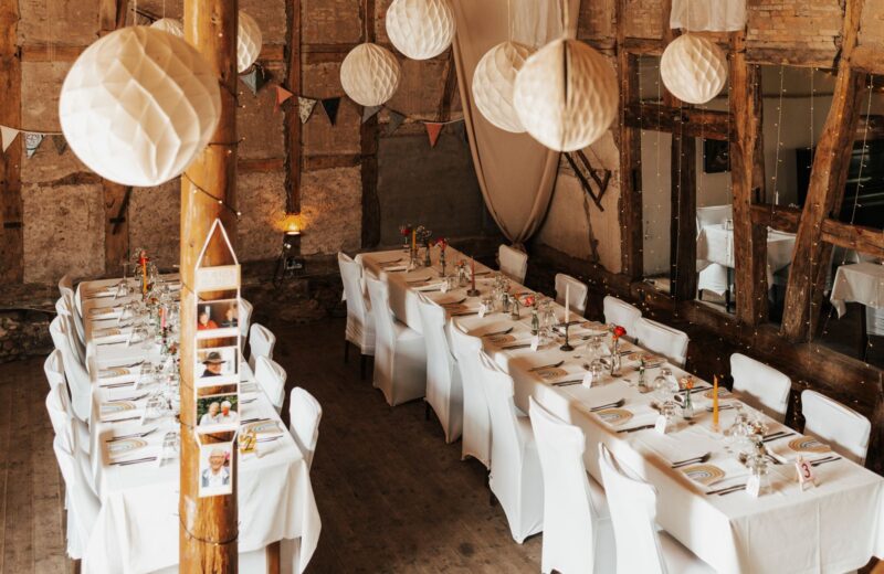 Long reception tables with white chairs under paper lanterns in a barn